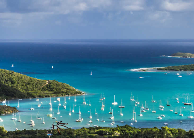 View of a scenic Caribbean bay with numerous sailboats on turquoise waters, surrounded by lush green islands under a partly cloudy sky.