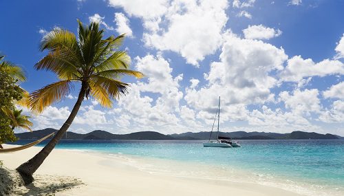 Tropical beach with palm tree and sailboat on turquoise water under blue sky with clouds.