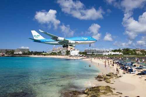 KLM airplane flying low over Maho Beach in Saint Martin, with tourists watching from the shore and a clear blue sky above.