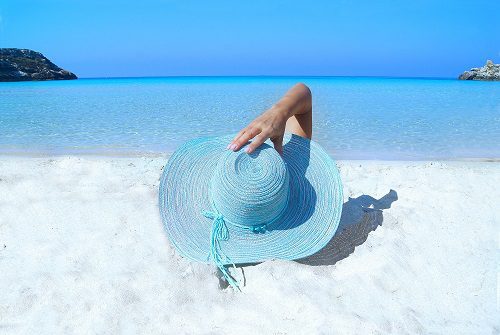 Person relaxing on a sandy beach near clear blue sea with a large sun hat.