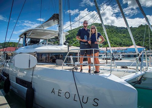 Couple standing on sailing yacht Aeolus at marina with green hillside and blue sky background.