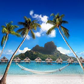 Tropical island paradise with palm trees, overwater bungalows, and a hammock on a sandy beach against a mountainous backdrop.