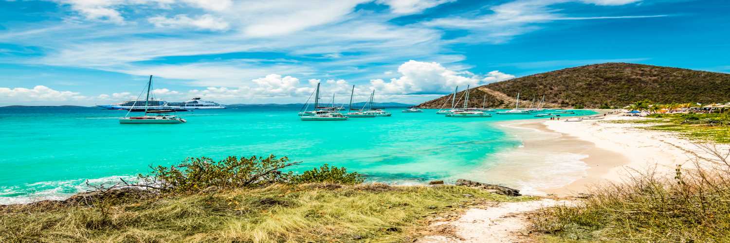 Tropical beach with turquoise water, sailboats anchored near the shore, and a hillside landscape under a blue sky.