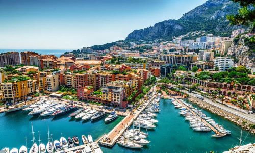 Aerial view of Monaco harbor with luxury yachts, waterfront buildings, and surrounding hills.