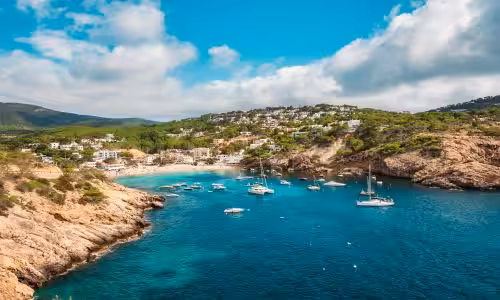 Scenic view of a coastal bay with turquoise waters, rocky cliffs, and sailboats anchored near a Mediterranean island, under a partly cloudy sky.