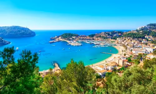 Scenic view of Port de Sóller in Mallorca with turquoise waters, vibrant coastal town, lush green hills, and clear blue sky.