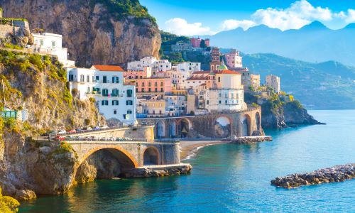 Scenic view of Amalfi Coast, Italy, featuring colorful cliffside buildings, arched bridge, and tranquil blue sea with mountainous backdrop.