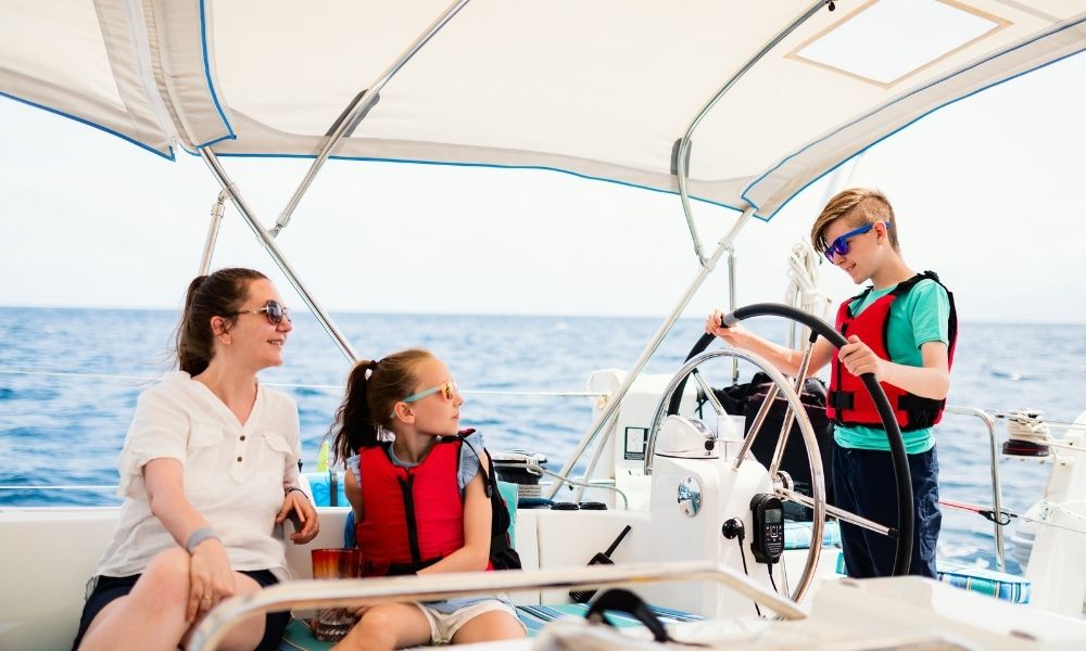 Family enjoying sailing on a sunny day, children wearing life vests and sunglasses, open sea background.