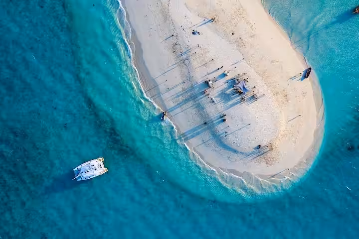Aerial view of a small sandy island with people gathered and a boat anchored nearby in clear turquoise water, ideal tropical destination.