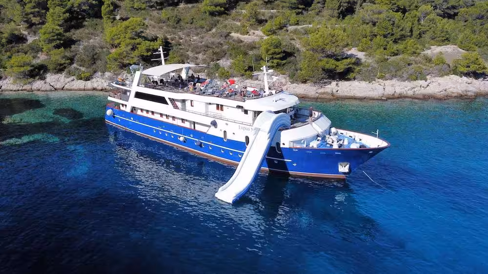 Luxury yacht with waterslide anchored near a scenic coastline with clear turquoise water and lush green trees.