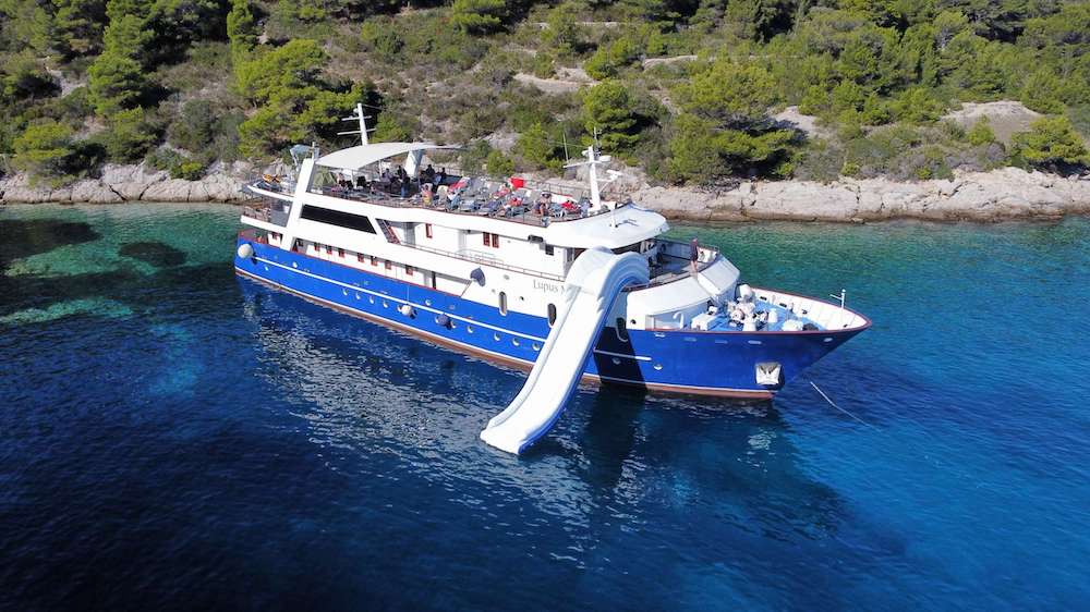 Luxury yacht with waterslide anchored near a scenic coastline with clear turquoise water and lush green trees.