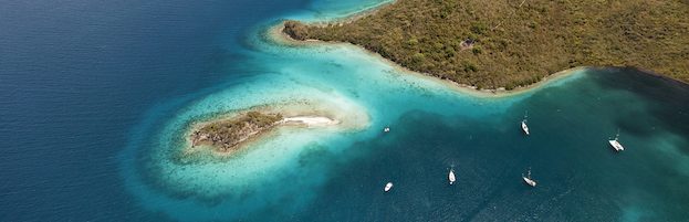 Aerial view of a turquoise tropical coastline with small island and anchored sailboats in clear blue waters.
