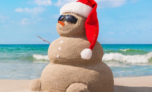 Sand snowman wearing sunglasses and a Santa hat on a sunny beach with ocean waves in the background.