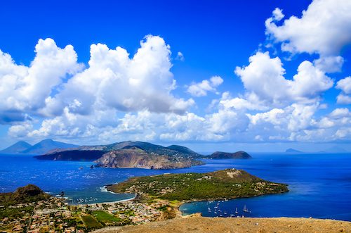 Scenic view of Lipari Island in Italy with lush green terrain, clear blue sea, and vibrant sky with fluffy clouds.