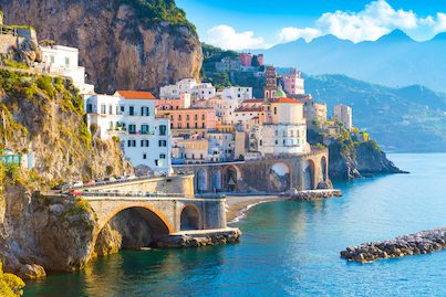 Scenic view of Amalfi Coast village with colorful houses and arched bridge over clear blue sea, mountain backdrop
