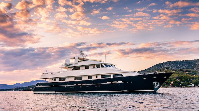Luxury yacht sailing on a calm sea during sunset with vibrant clouds and distant hills in the background.