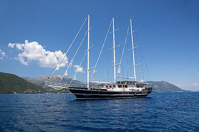 Luxury sailing yacht with multiple masts cruising on clear blue waters surrounded by scenic mountain landscape under a clear sky.
