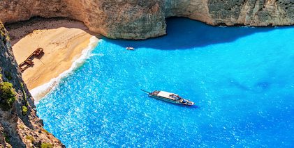 Navagio Beach in Zakynthos, Greece with clear turquoise water, a shipwreck on the sandy shore, and a boat floating nearby.