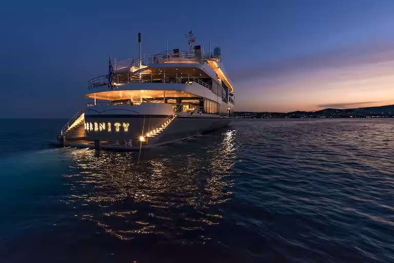 Luxury yacht Serenity illuminated at dusk, cruising on serene waters with a beautiful sunset in the background.