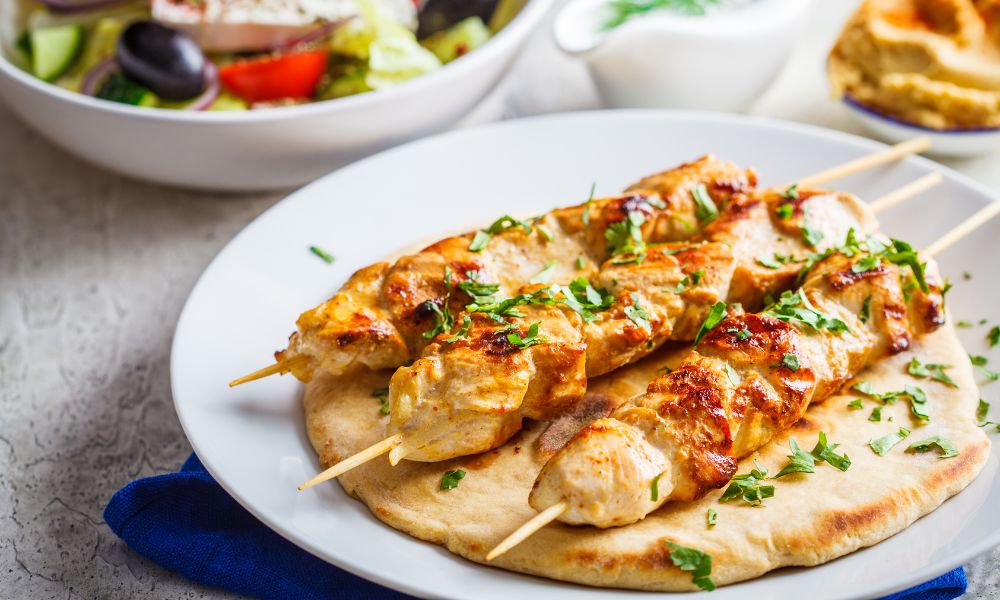 Grilled chicken skewers garnished with herbs on pita bread, served with a Greek salad and hummus on a rustic table setting.