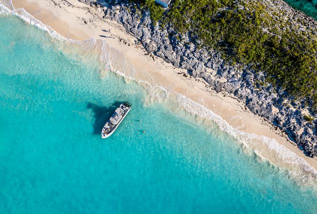 Aerial view of a boat anchored near a pristine sandy beach with clear turquoise water and lush greenery on a sunny day.