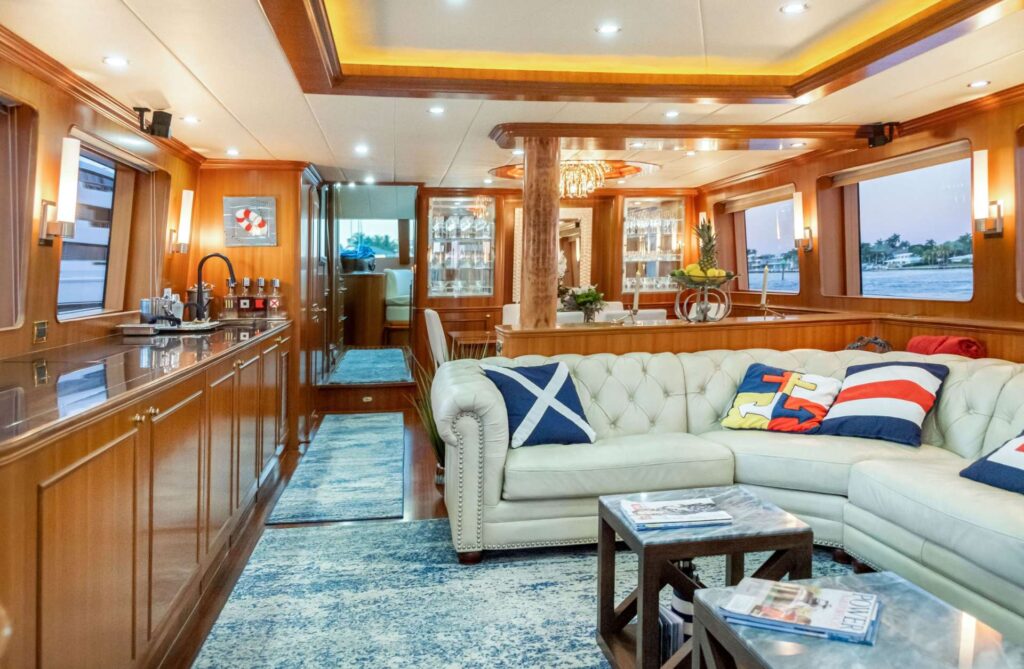 Luxurious yacht interior featuring polished wood finishes, a cozy white leather sectional with nautical-themed pillows, a sleek bar area, and large windows offering scenic ocean views.