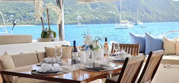Luxury yacht dining setup overlooking serene ocean with mountains, featuring elegant table setting including wine bottles, glasses, and floral centerpiece, with plush seating and scenic sailboats in the background.