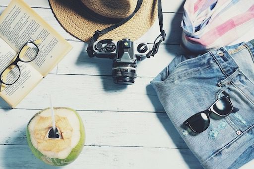 Travel essentials including sunglasses, camera, straw hat, open book, denim shorts, scarf, and a fresh coconut drink on a white wooden table.