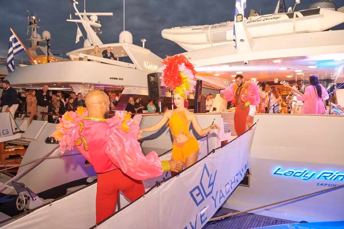 Dancers in colorful costumes perform on a luxury yacht during an evening event, surrounded by an enthusiastic crowd.