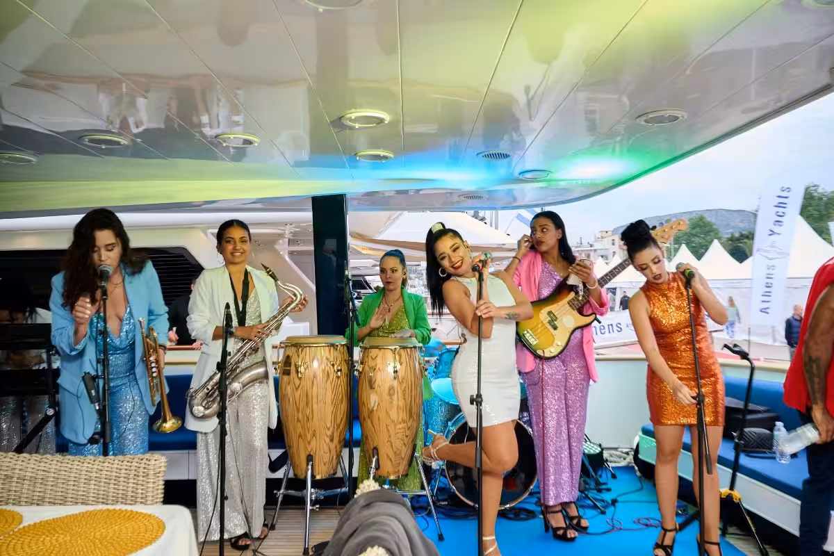 All-female band performing energetically on a yacht, featuring brass and percussion instruments, vibrant outfits, and an enthusiastic singer, set against a backdrop of luxury yachts and festive atmosphere.