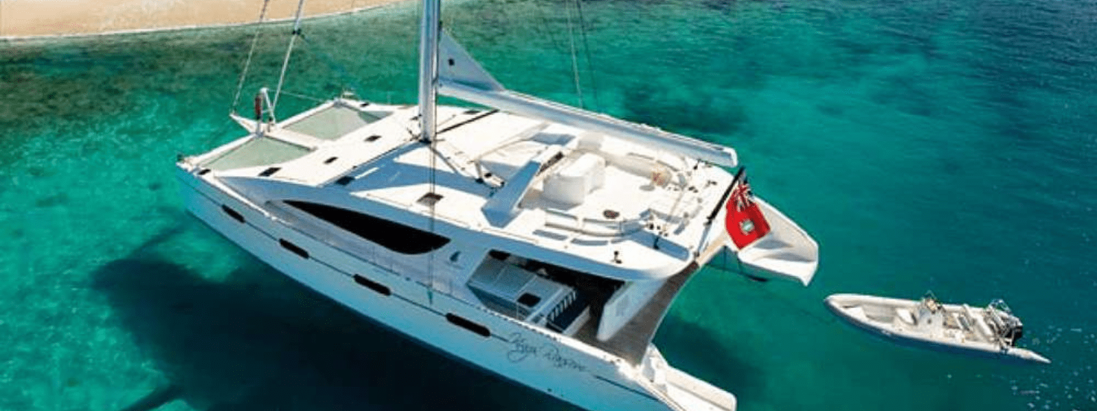 Luxury catamaran anchored in crystal-clear turquoise waters with a small dinghy attached, near a sandy beach.