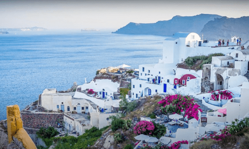 Scenic view of Santorini coastline with whitewashed buildings and vibrant bougainvillea, overlooking the Aegean Sea and distant islands.