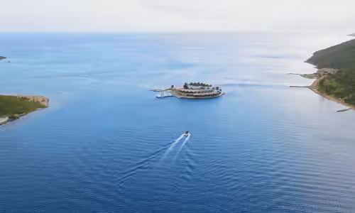Aerial view of an idyllic island resort surrounded by vibrant blue ocean waters with a boat leaving a white trail, showcasing tropical getaway scenery.