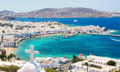 Aerial view of Mykonos town with whitewashed buildings, turquoise waters, and a coastline dotted with boats and windmills on a sunny day.