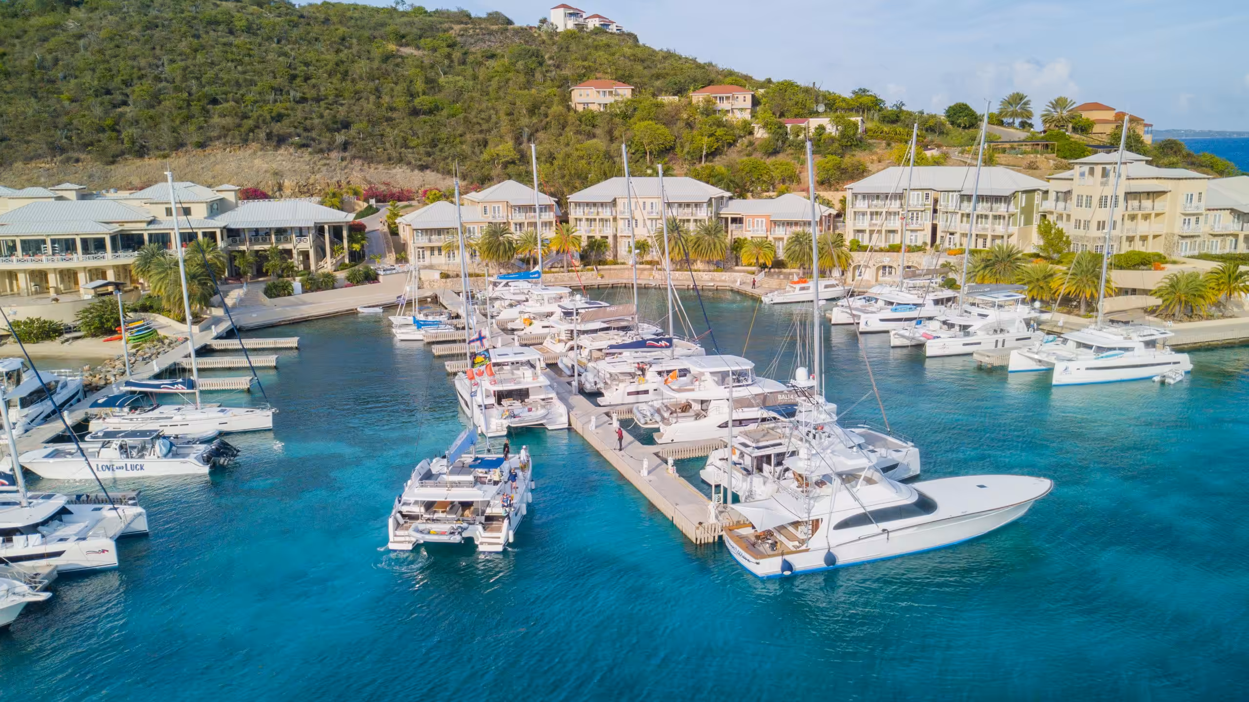 Marina with luxury yachts docked in turquoise waters, surrounded by tropical villas and lush green hillside.