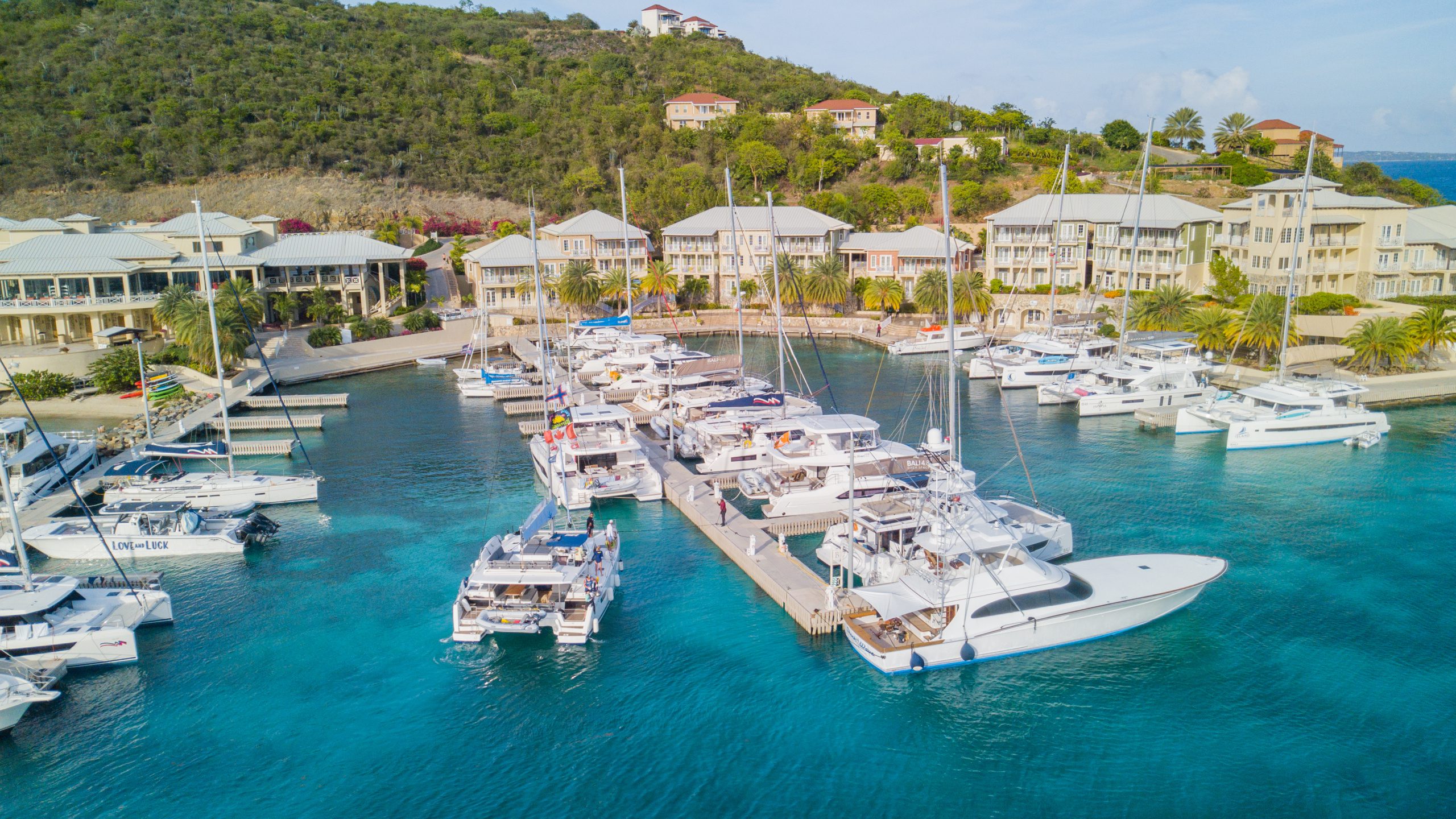 Marina with luxury yachts docked in turquoise waters, surrounded by tropical villas and lush green hillside.